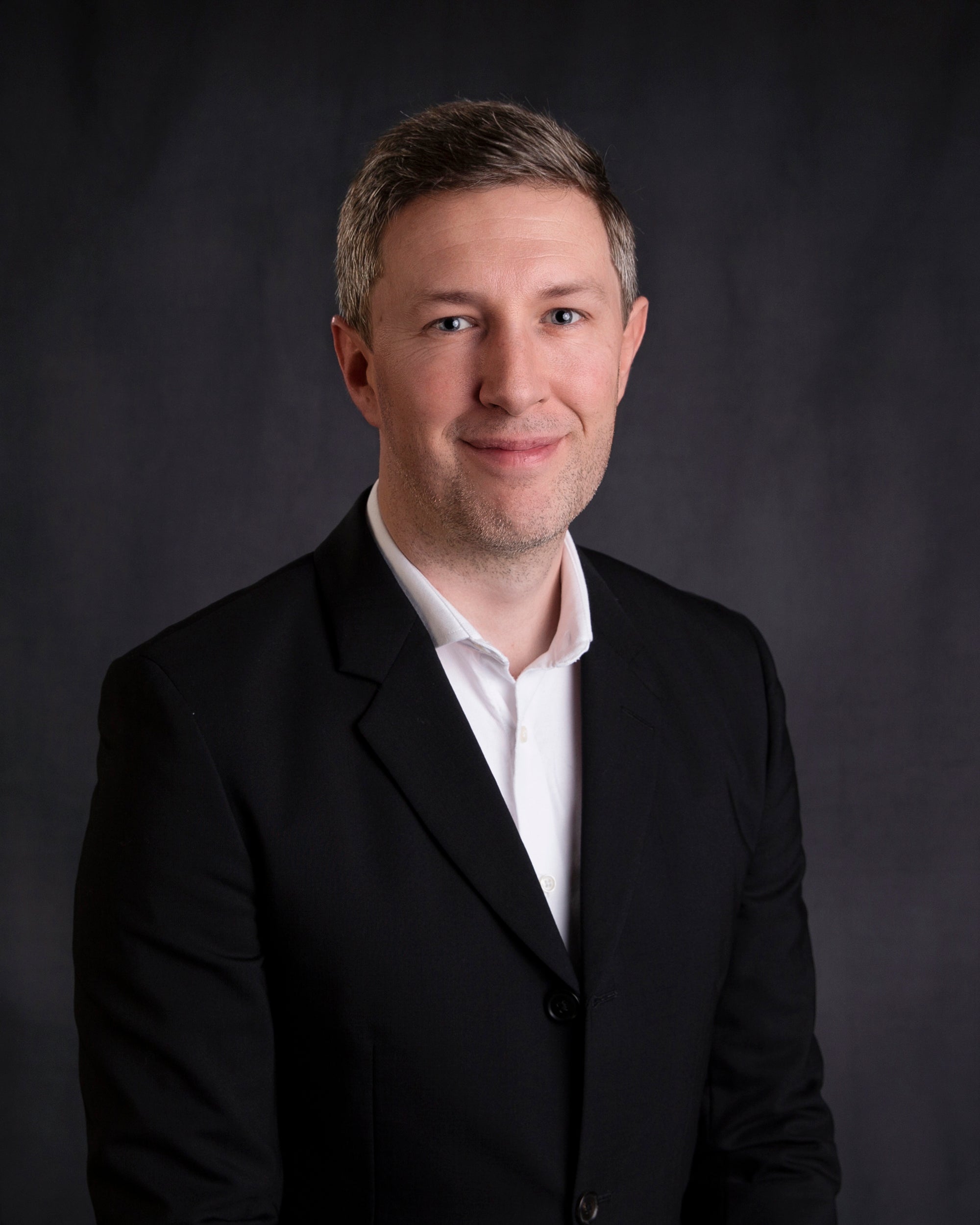 Mark Whitney wearing a black suit and white shirt against a dark background